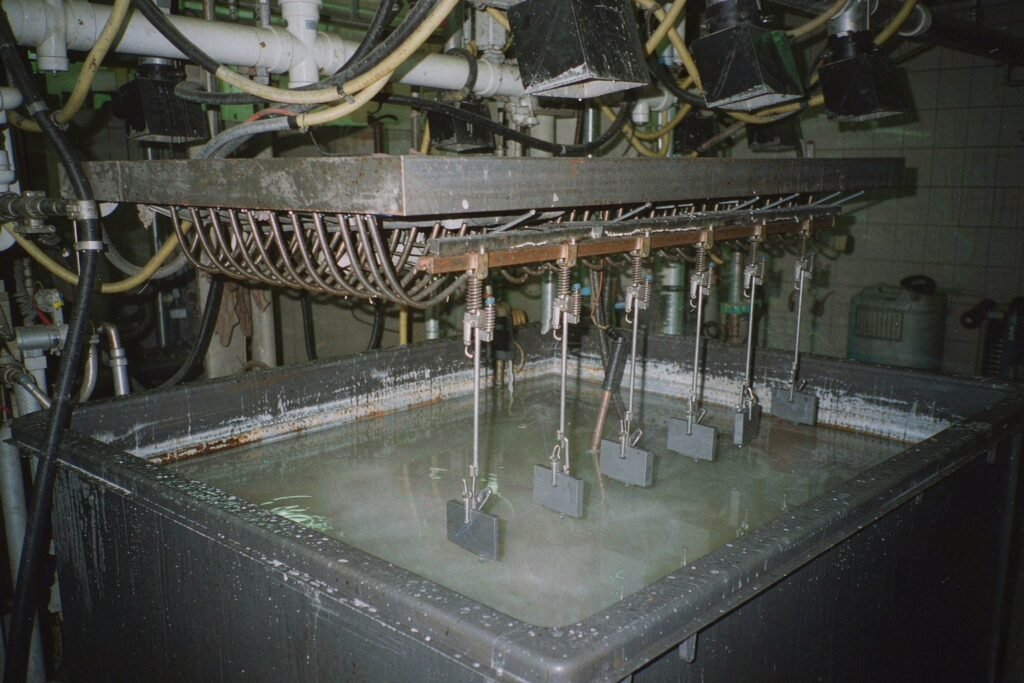 Machined aluminium parts held on spring-loaded titanium fingers above a sulphuric acid anodising tank, copper busbar along the rim and the still surface of the electrolyte below in a surface treatment workshop