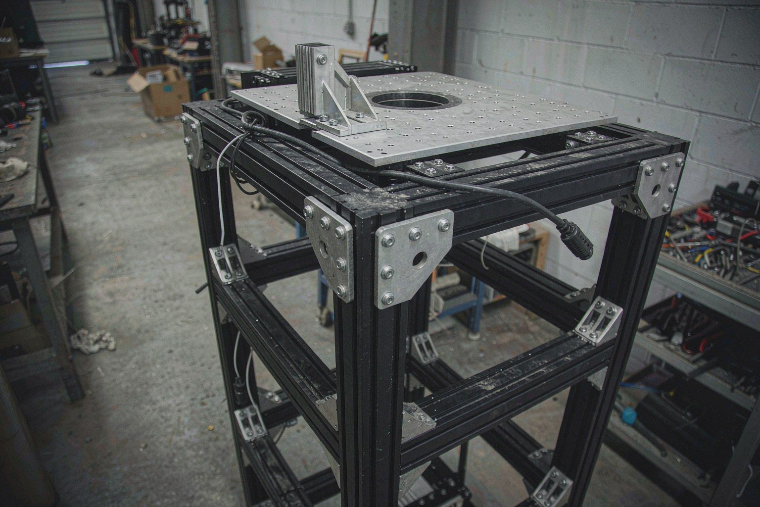 A black-anodized aluminum T-slot automation frame with cast corner brackets, carrying a machined aluminum mounting plate with a central motor bore, a ring of clearance holes and a ribbed L-bracket with triangular gussets