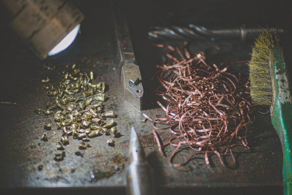 Short broken brass chips piled beside a tangle of long copper swarf ribbon, with a worn carbide turning tool and a chip brush on the machine table