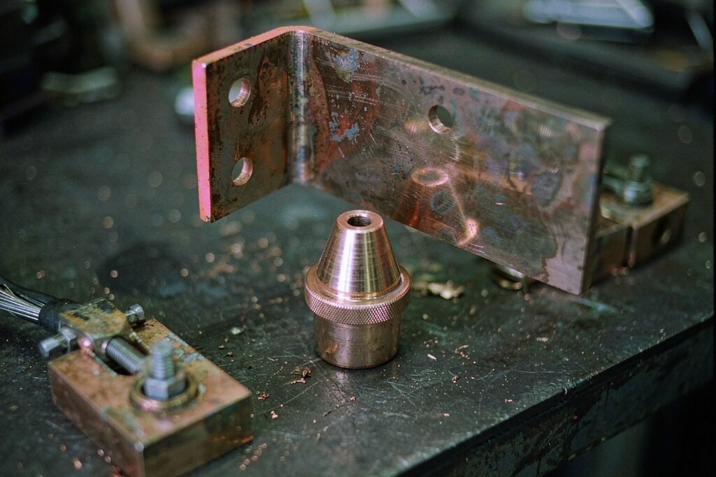 Copper conductive parts on a machine shop bench: a busbar bent at ninety degrees with mounting holes, a tapered welding nozzle with a knurled collar and a ground block