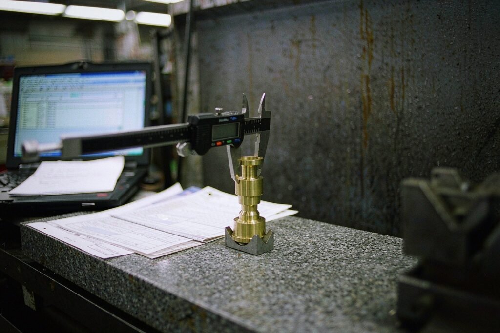 A machined brass fitting held in a V-block on a granite surface plate while a digital caliper measures it, with a blank inspection report beside it