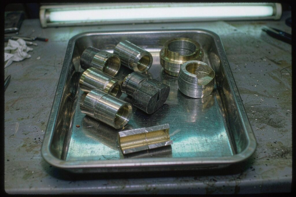 Tin-plated and nickel-plated brass turned parts in a steel tray, one part sectioned to show the plating layer over the brass core