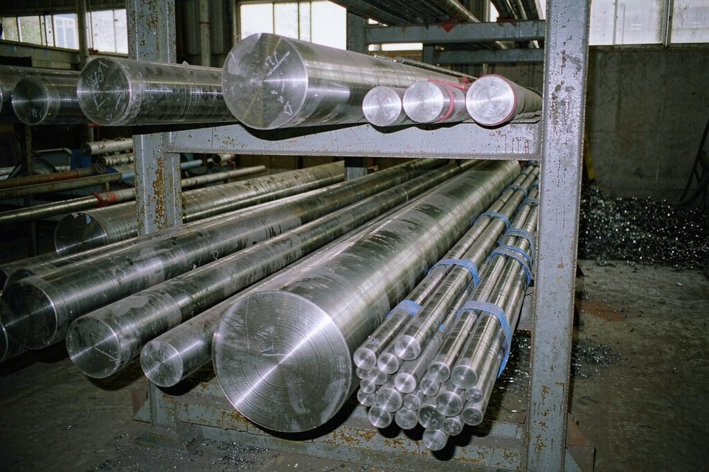 Stainless steel round bar stock stacked in a cantilever rack, bright cold-finished surfaces, cut ends showing fine concentric turned faces and a bundle still in blue protective sleeving