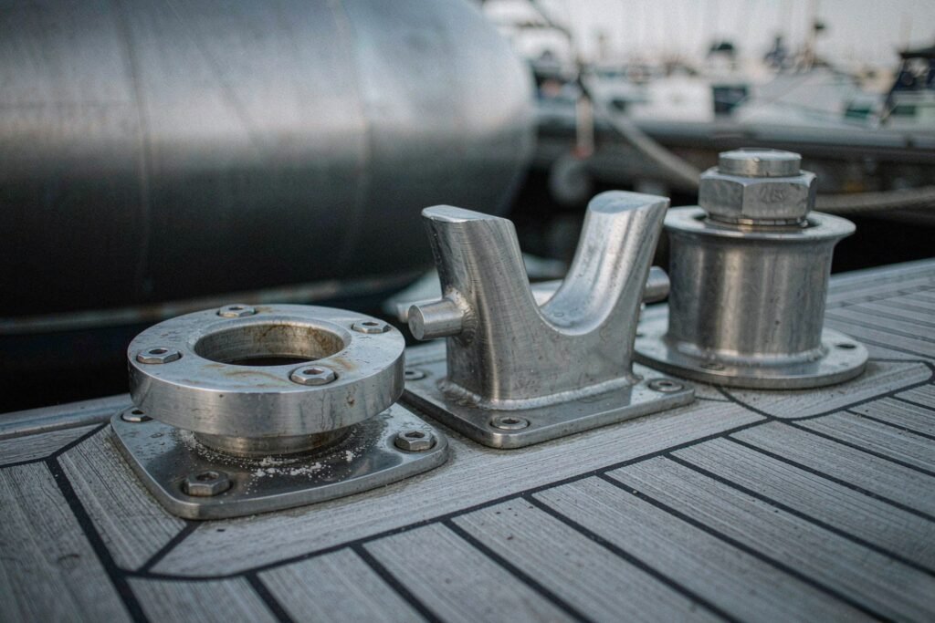 Stainless steel marine deck hardware on a weathered teak deck, a bow eye fitting with four countersunk bolts and tea-staining beginning in a shielded crevice