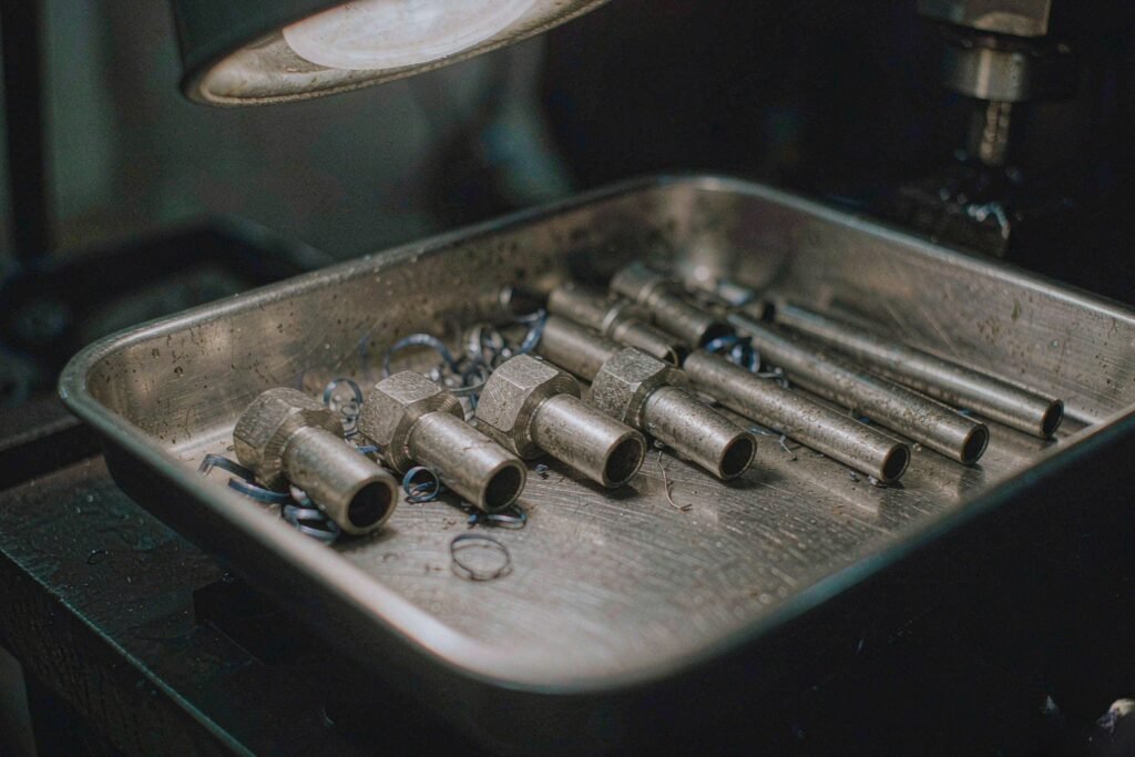 A tray of CNC turned stainless steel parts - hex-bodied hose fittings, stepped pins with shoulder fillets, a grooved valve spool body and thin-walled sleeves - with curled stainless swarf