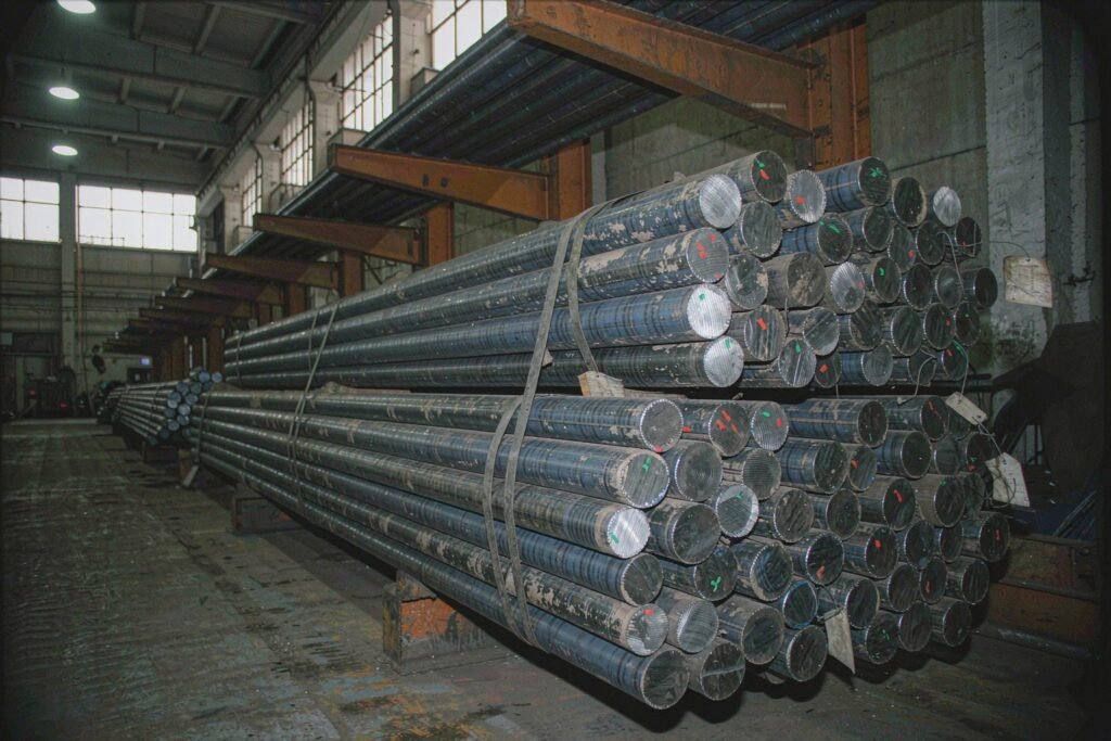 Bundles of hot-rolled carbon steel round bar in warehouse racks, dark blue-grey mill scale surfaces, sawn circular ends showing bright cut faces, steel banding and wired paper mill tags