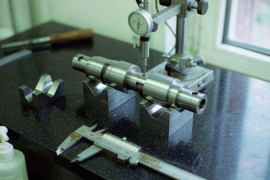 A stepped steel shaft supported in V-blocks on a granite surface plate while a dial indicator measures a journal, with a vernier caliper and a blank inspection form