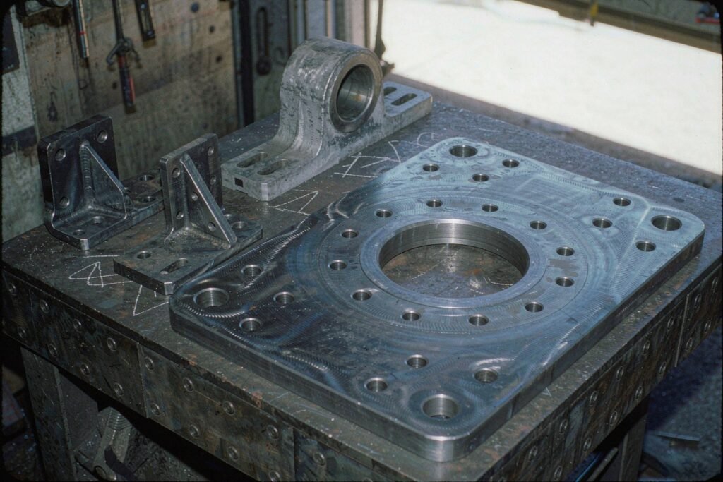 Machined carbon steel mechanical structures on a fabrication bench: mounting plates with a large central bore and a ring of bolt holes, gusseted L brackets and a pillow block bearing housing