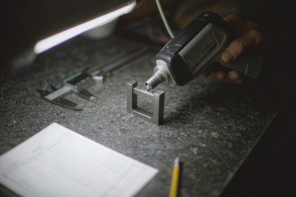 A titanium bracket being checked with a handheld alloy analyser on a granite surface plate beside a printed inspection report and a caliper