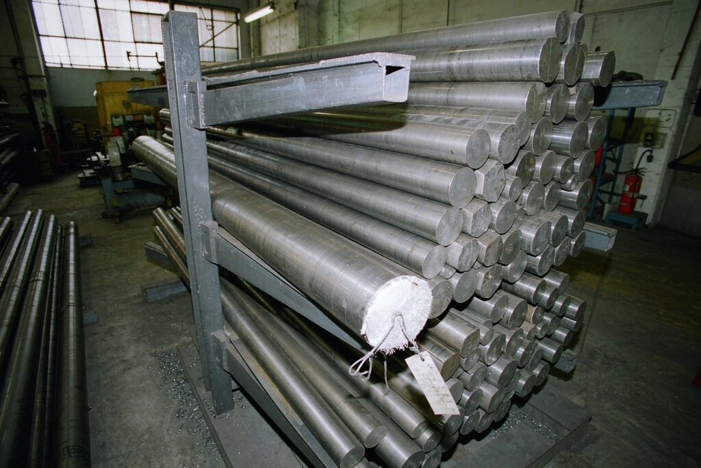 Titanium round bar and hex bar stock racked in a machine shop, one bar end showing a bright bandsaw cut face and a tied-on mill tag