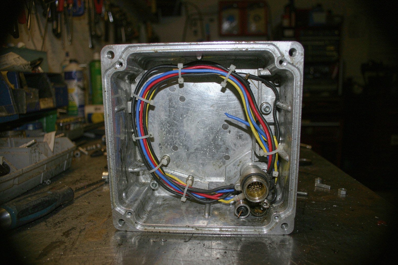 A cable harness routed along the inside wall of an open machined aluminium enclosure, eight insulated wires held by nylon cable ties on tie-down studs, ending in a circular metal-shell connector with a coupling ring
