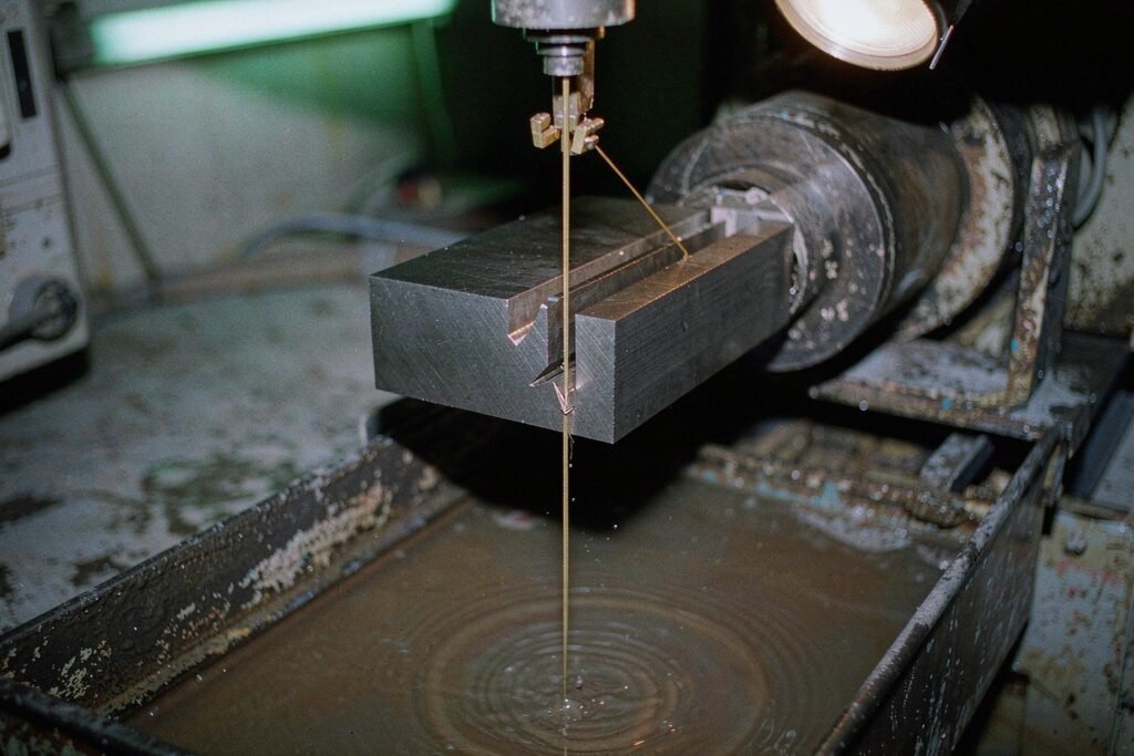 Wire EDM cutting a narrow slot in a hardened steel block, a thin brass wire running through the workpiece with sparks at the cut and dielectric fluid flushing over it