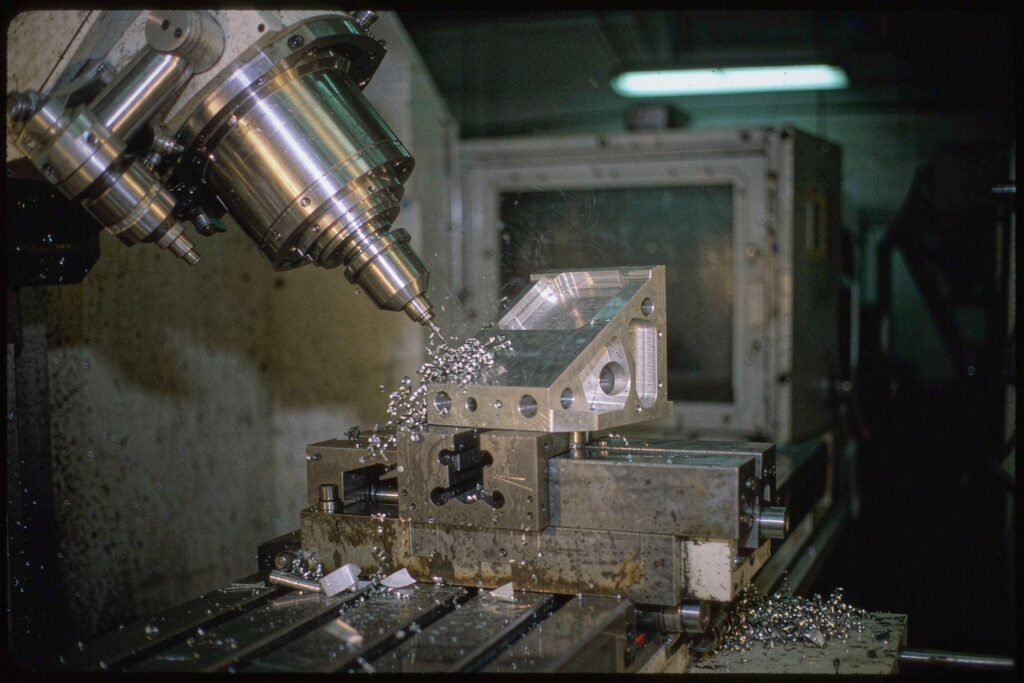 Five-axis machining center cutting an aluminium bracket at a compound angle, chips flying, coolant on the table and the machine enclosure out of focus behind