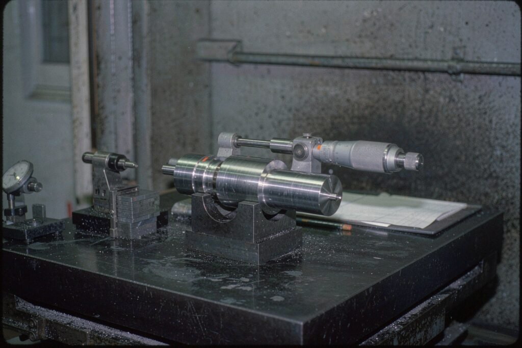 A precision stepped shaft held in a V-block on a granite surface plate, measured with an outside micrometer across a ground diameter, gauge blocks and a dial test indicator beside it