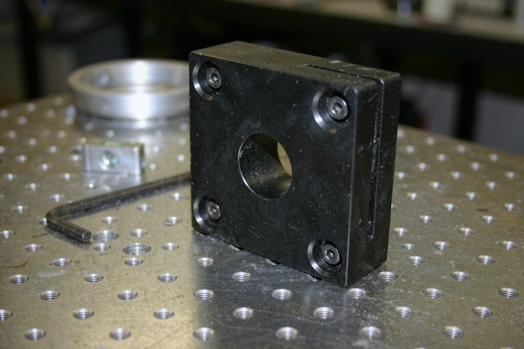 An anodised aluminium optical mounting bracket with a bored through-hole and counterbored mounting holes, bolted to a stainless optical breadboard with a threaded hole grid