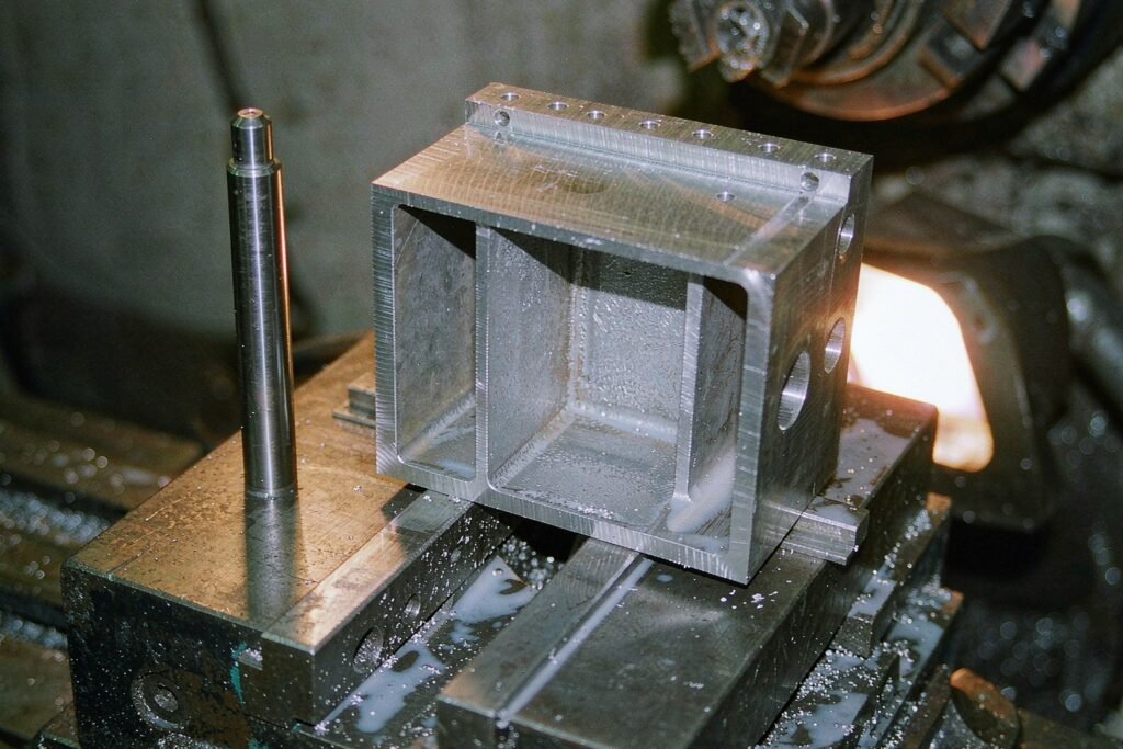 A thin-walled aluminium part with a deep milled cavity on a machine table, 2 mm rib walls, a ring of bolt holes along the flange and a long-reach end mill standing beside it for scale
