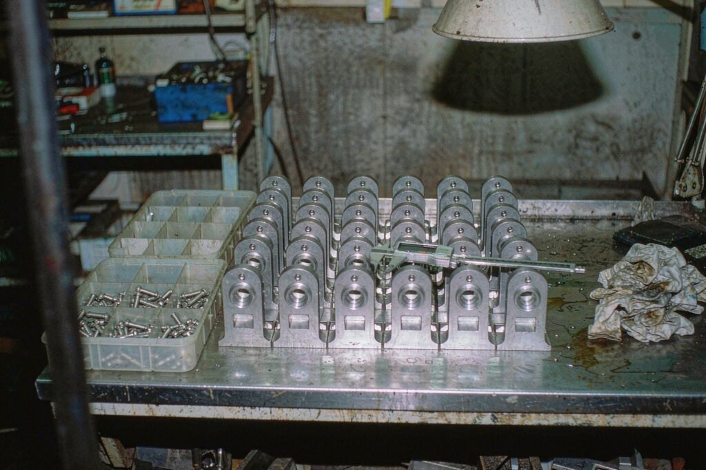 About twenty identical small machined aluminium brackets arranged in rows on a steel workbench, a tray of socket head screws and a caliper lying beside them