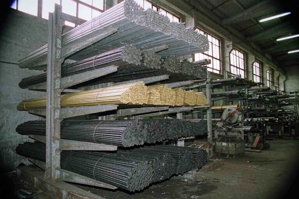A cantilever stock rack holding bundles of bar stock: bright aluminium round bar, dark steel hex bar, yellow brass round bar and black plastic rod, cut ends showing saw marks