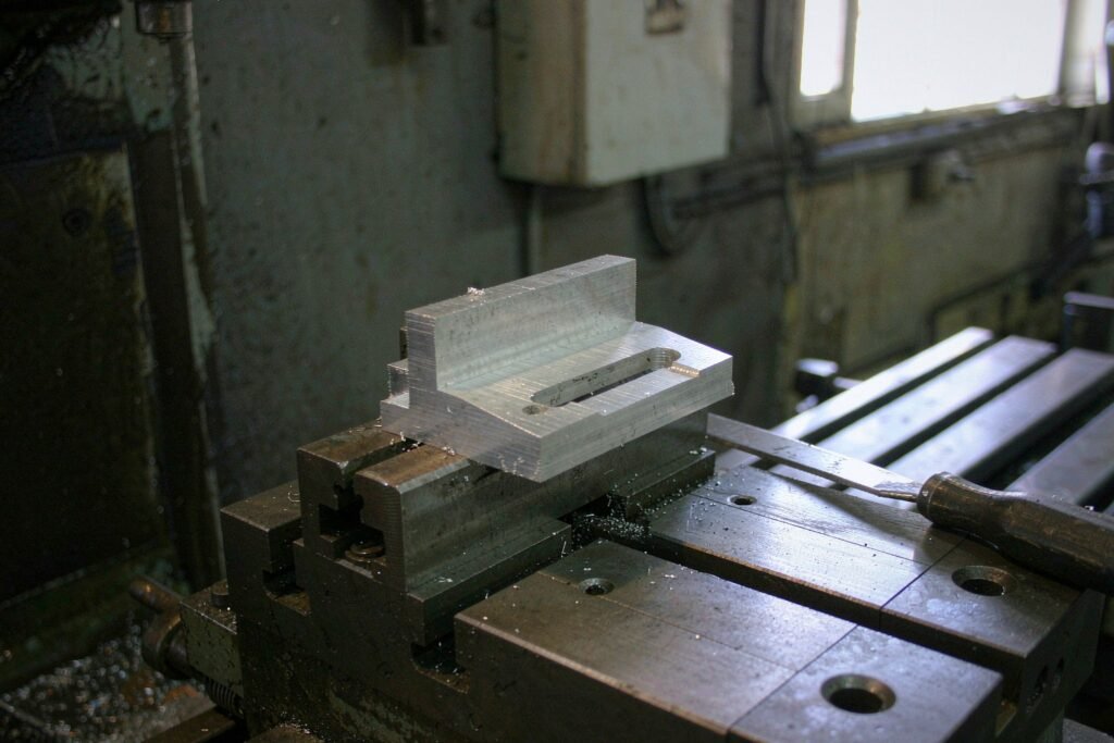 A single aluminium prototype bracket still clamped in a machine vice, stepped milled faces, a slotted hole and a chamfered edge, a hand deburring file lying beside it on an otherwise empty worktable