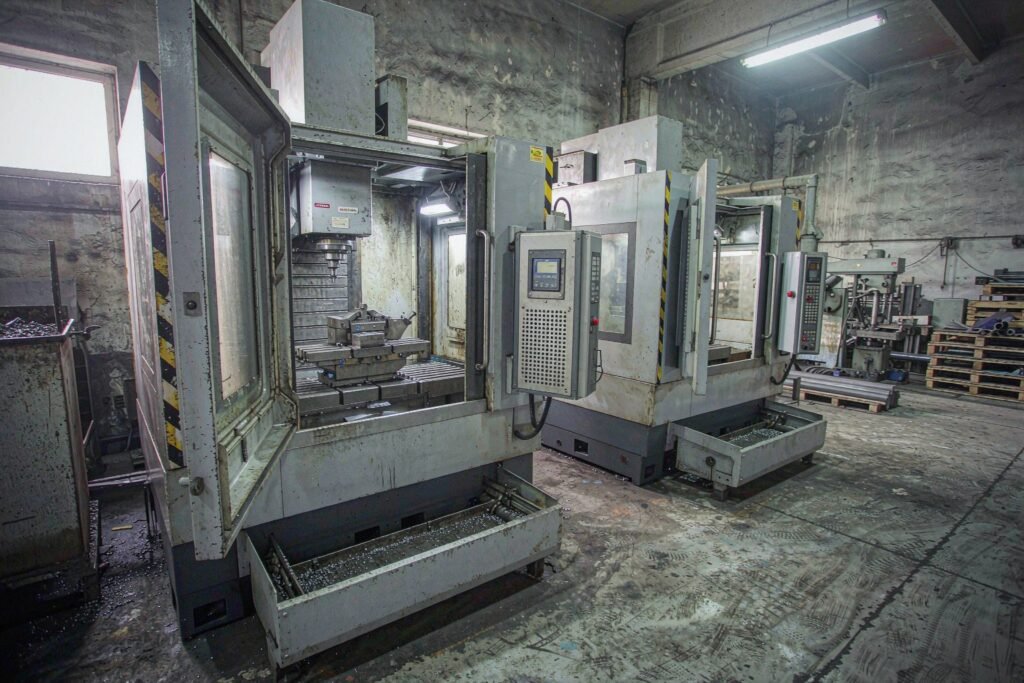 An aisle between two CNC machining centres in a working machine shop, one machine door open on a T-slot table with a part in the vice, chip conveyors, control pendants and a pallet of stock in the background