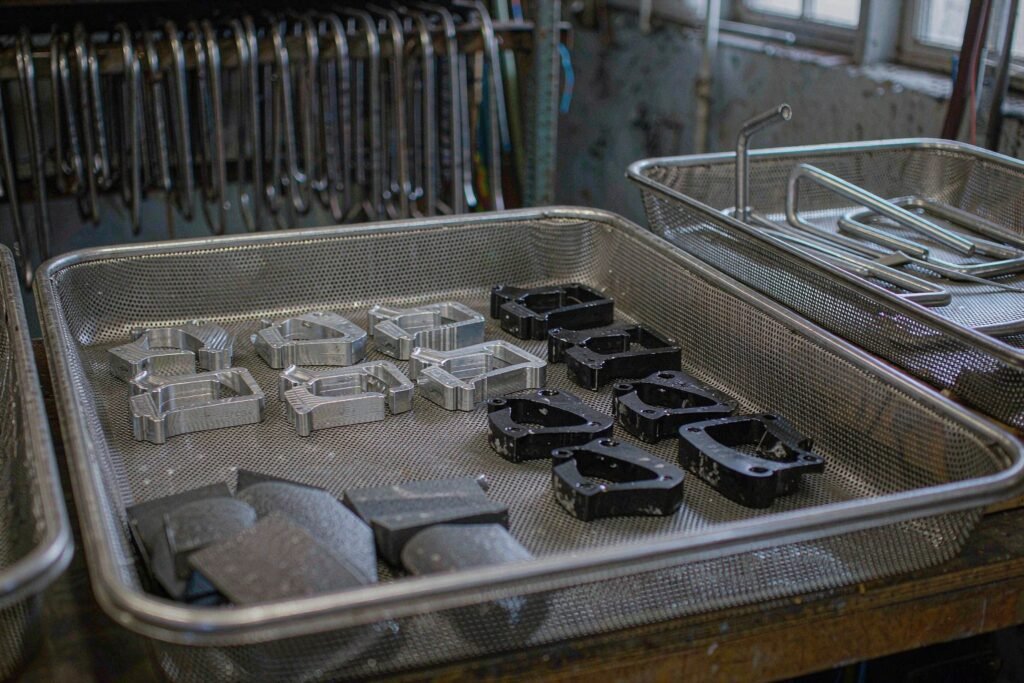 A steel mesh tray of small aluminium parts after surface treatment, half bare machined aluminium with visible tool marks and half matte black anodised, with bead-blasted stainless parts in one corner