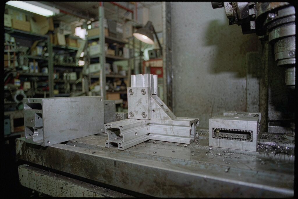 Automation equipment components on a machine table: an aluminium mounting bracket with slotted holes, an extruded frame corner joined by a gusset plate, and a linear rail carriage block with a short length of profiled rail