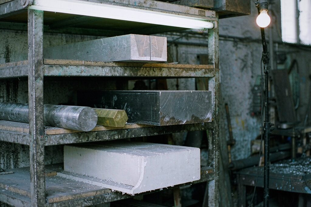 Raw material billets on a steel rack: a bright aluminium block, a dark steel block with mill scale, a stainless steel round bar, a yellow brass hex bar and a white engineering plastic block