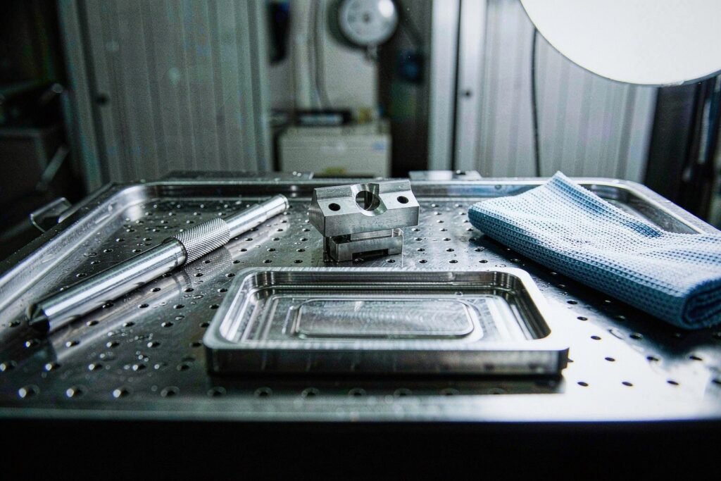 Medical device components on a perforated stainless tray: a thin stainless instrument housing shell with a milled recess, a slender knurled instrument shaft and a small titanium sensor mount bracket