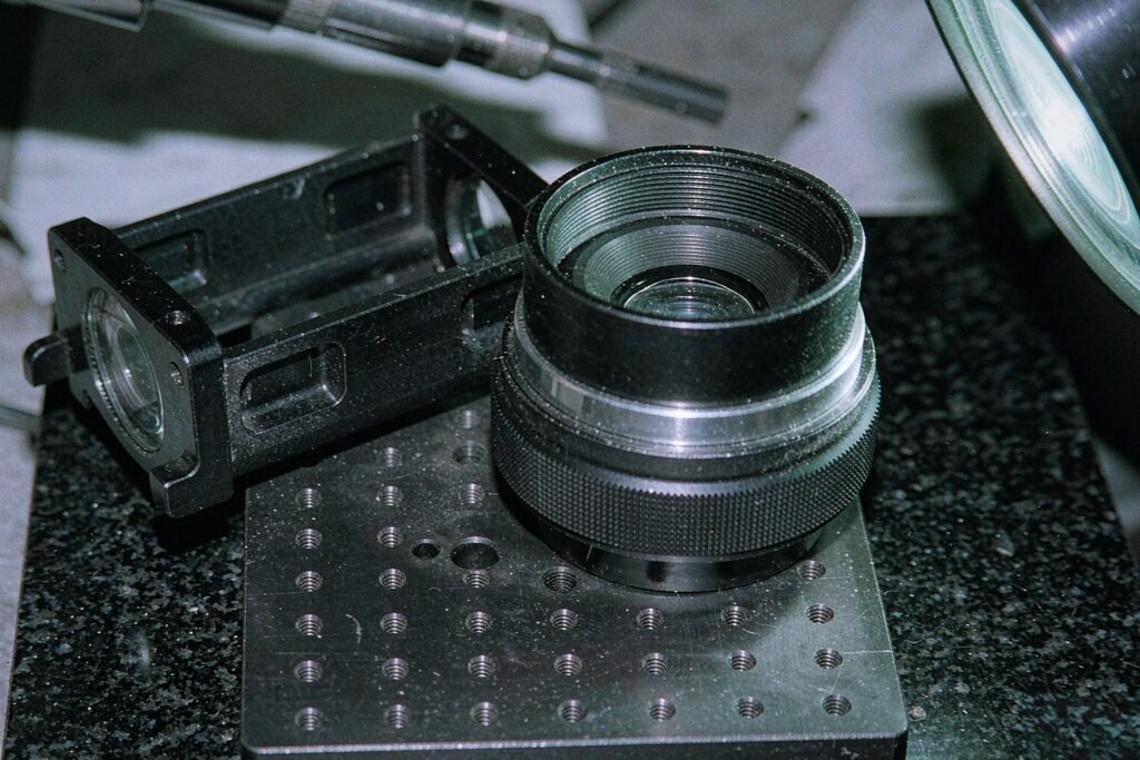 Optical system parts on a granite plate: an aluminium lens housing barrel with a threaded bore and knurled focusing ring, a black anodised alignment fixture plate and an optical mounting frame with milled lightening pockets