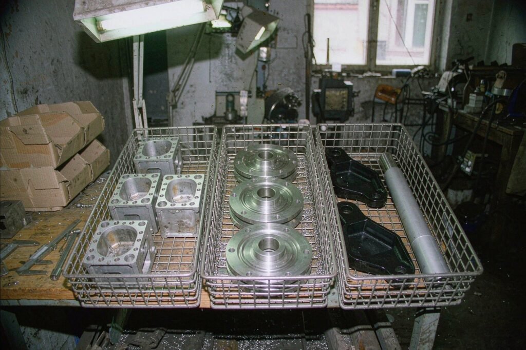 Three steel mesh trays of finished CNC parts on a bench, one with aluminium housings, one with stainless steel flanges and one with black anodised brackets and a bead-blasted shaft, a caliper lying across the bench