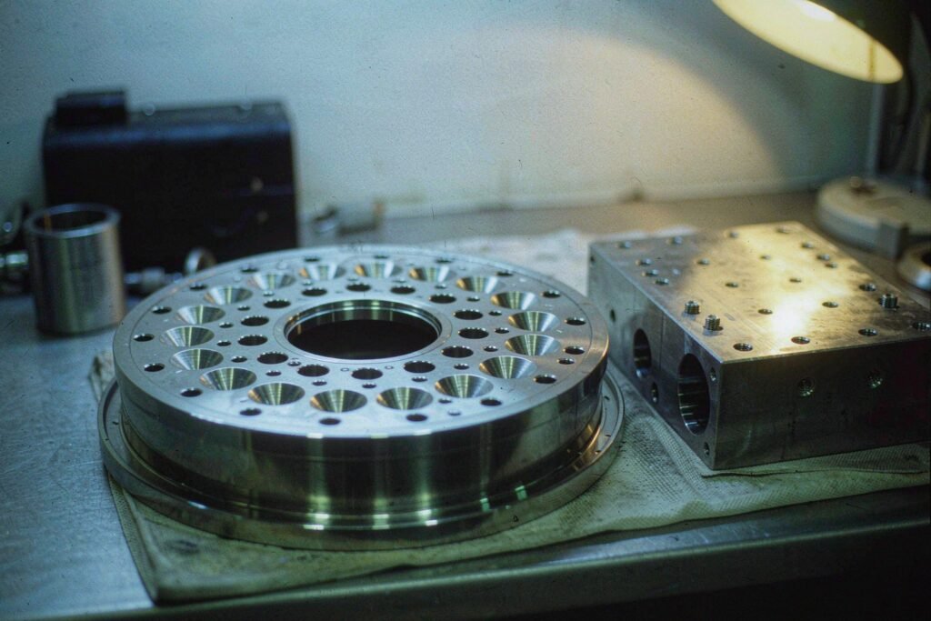 Semiconductor vacuum components on a bench: a circular stainless flange with a ring of bolt holes and a machined knife-edge sealing ring around the bore, beside a rectangular vacuum manifold block with bored gas passages