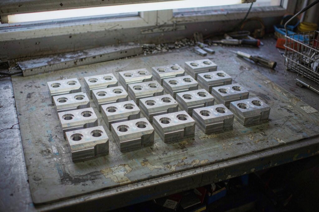 A finished low-volume batch of machined aluminium parts laid out in neat rows on a steel bench, raw milled finish with visible tool marks, a steel rule and deburring tool beside them