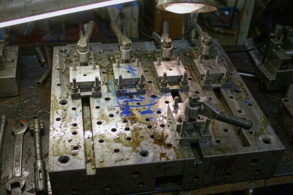 A dedicated multi-station machining fixture: thick steel base plate with T-slots, four identical parts clamped side by side against hardened locating pins by swing clamps, one empty station with the clamp open