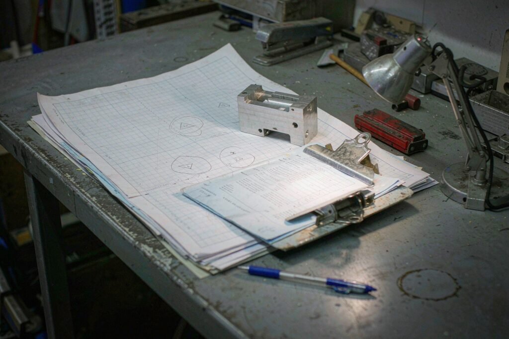 A job traveller and revision-control station: printed engineering drawings marked with revision clouds, a clipboard traveller sheet with handwritten ticks, a revision stamp and the matching machined part on top of the drawing