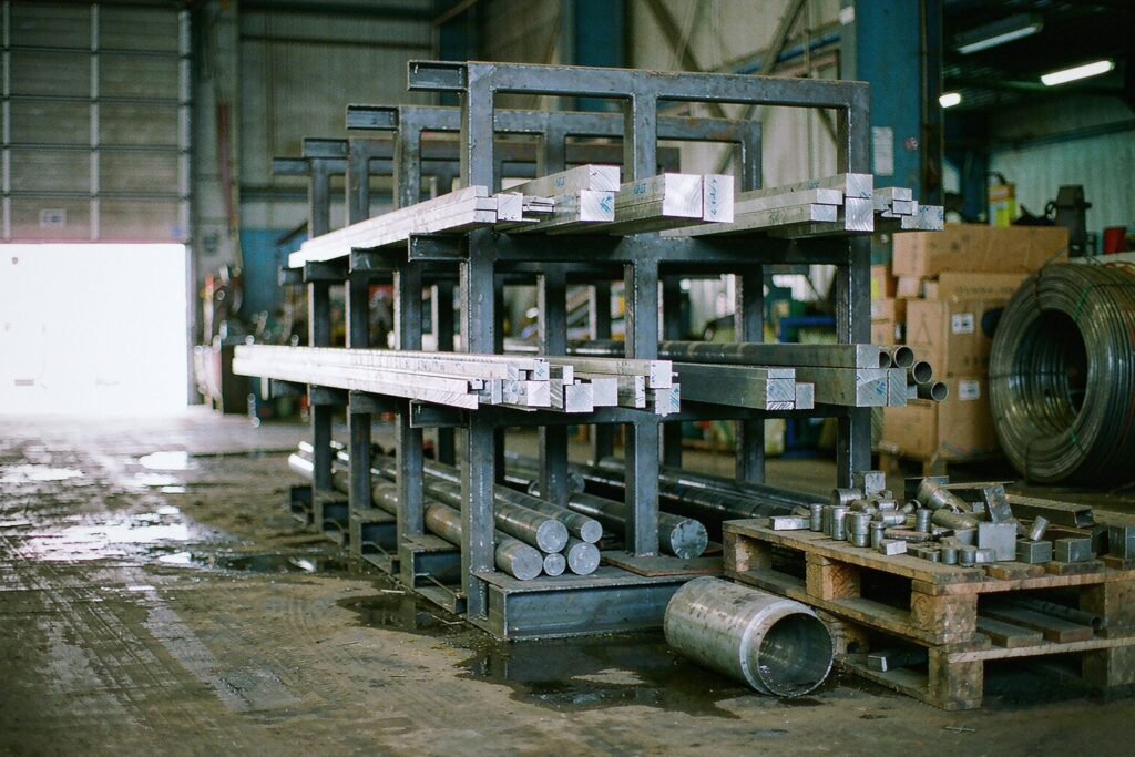 Raw material stock for a small batch: a cantilever rack of aluminium rectangular and round bar with bright saw-cut ends, and a pallet of offcuts on a grimy warehouse floor