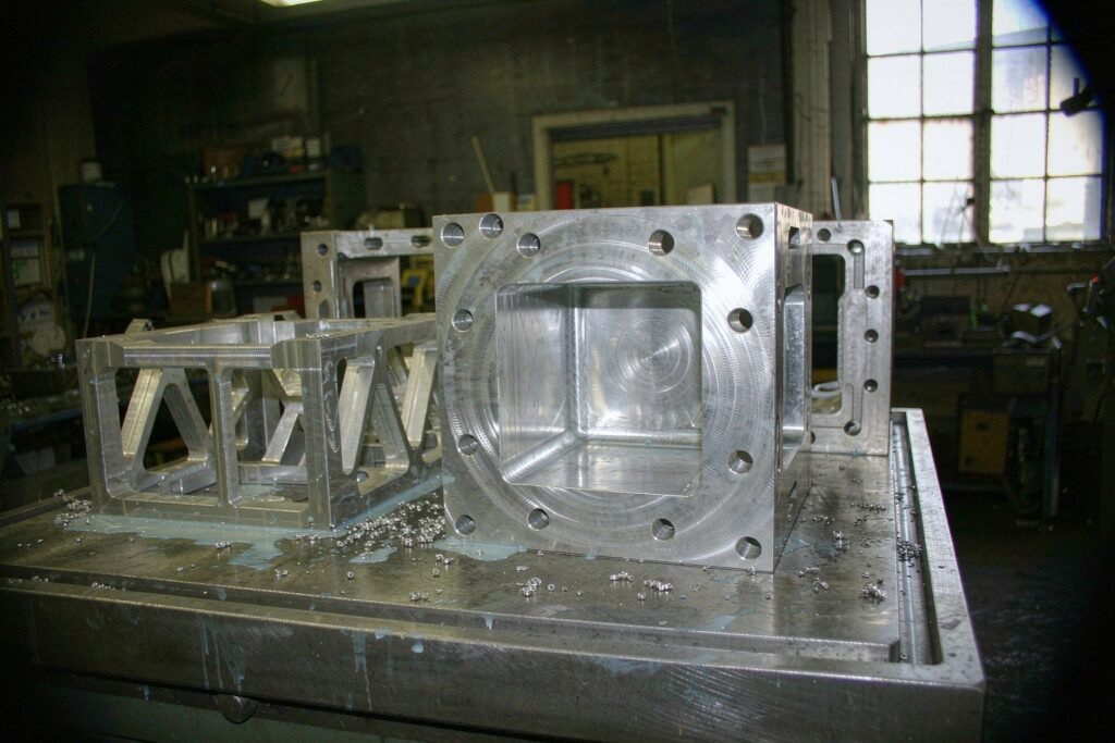 Machined aluminium parts on a steel bench: a 6061 housing with a milled cavity and a ring of bolt holes, a thin-walled frame with lightening pockets and an extruded mounting bracket with slotted holes