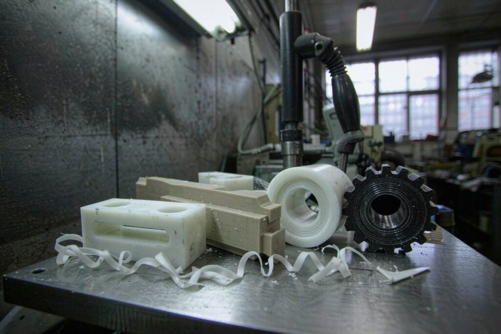 Engineering plastic machined parts on a steel table: a white POM sliding block with a milled slot, a beige PEEK spacer, a white PTFE bushing with turning rings and a black nylon gear blank with ribbony swarf