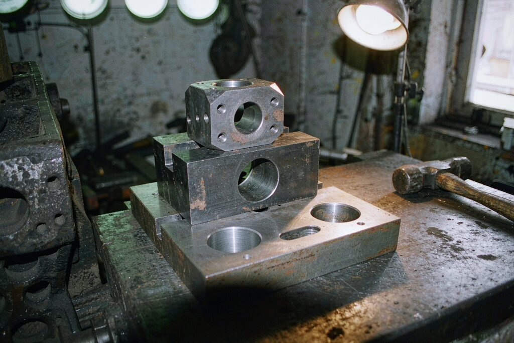 Carbon and alloy steel machined parts on a workbench: a thick C45 base plate with bored holes, a black-oxide load-bearing block with a keyway and a hex coupling body with radial tapped holes