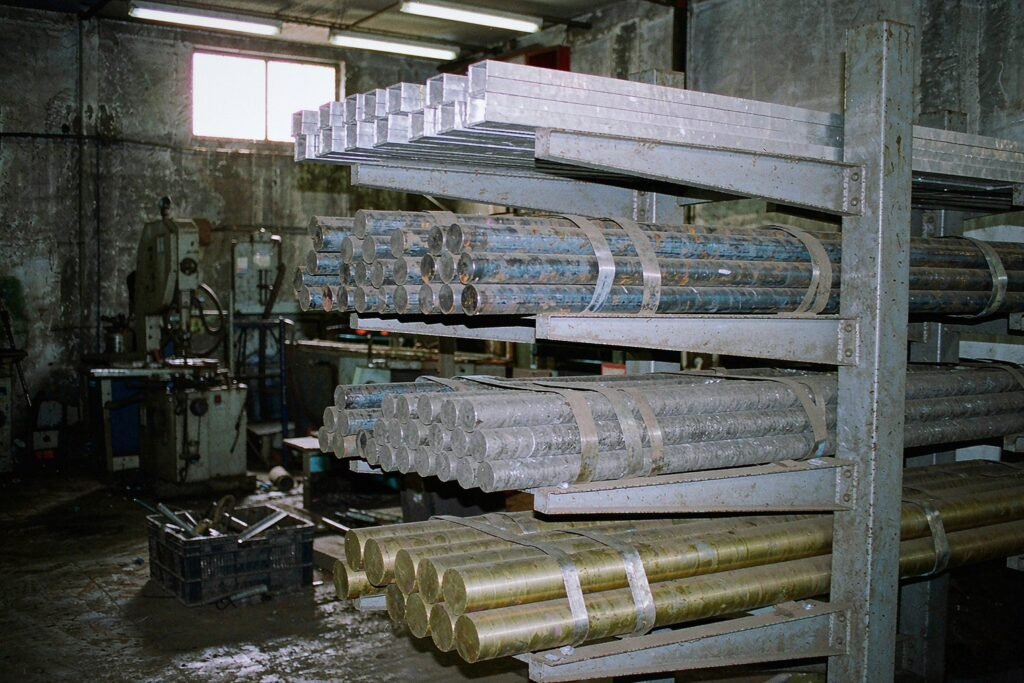 Raw material bar stock in a cantilever rack: bright aluminium blocks, dark mild steel round bar with mill scale, dull stainless steel bar and yellow brass hex bar, a band saw and offcuts bin behind