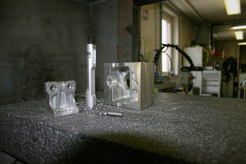 Titanium machined parts on a granite plate: a Ti-6Al-4V bracket with thin ribbed walls, a slender stepped medical implant trial part with a bead-blasted surface and a structural block with lightening pockets