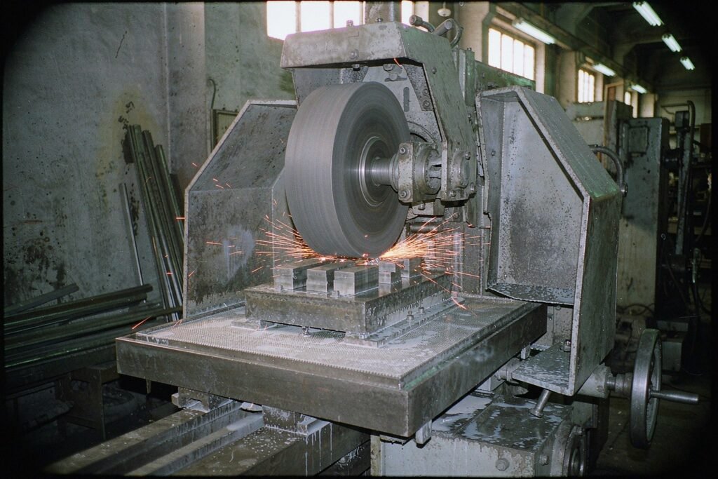 A precision surface grinder throwing sparks off hardened steel blocks on an electromagnetic chuck, whitish grinding sludge spread across the machine table