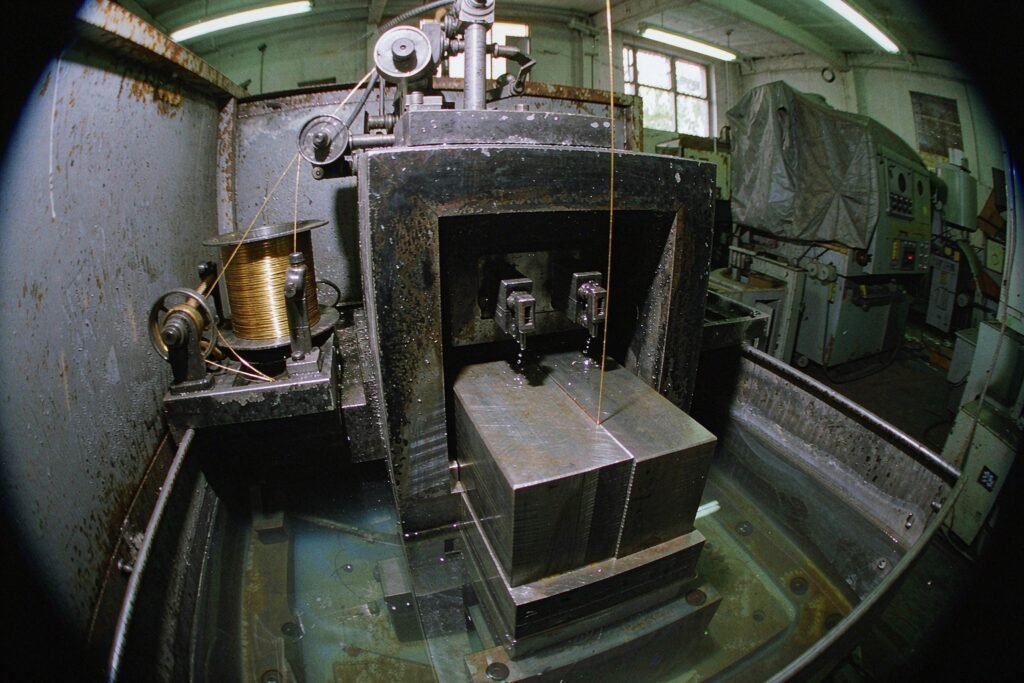 A wire EDM machine cutting submerged, the brass wire running through a steel plate in a tank of dielectric fluid beside the wire spool and take-up reel