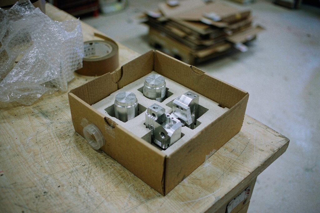 Prototype parts packed for dispatch in an open carton with die-cut foam inserts, bubble wrap, packing tape and a packing list on a wooden bench in a dispatch bay