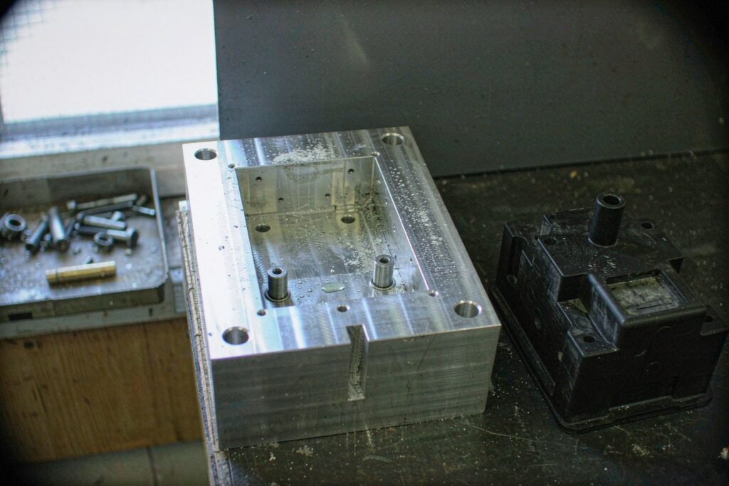 Soft bridge tooling on a bench: a thick aluminium mould plate with a milled rectangular cavity, two hardened locating pins, four clearance bolt holes and a matching moulded part beside it