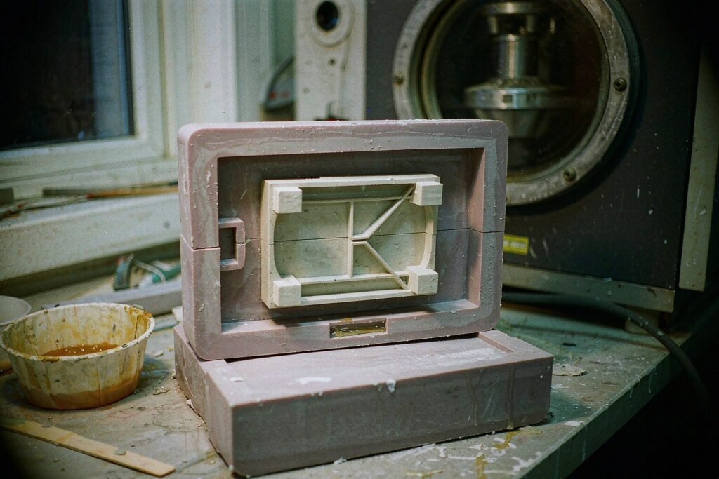 A two-part silicone casting mould opened flat on a bench with a cast polyurethane prototype part sitting in the cavity, a mixing cup of resin and a vacuum casting machine behind